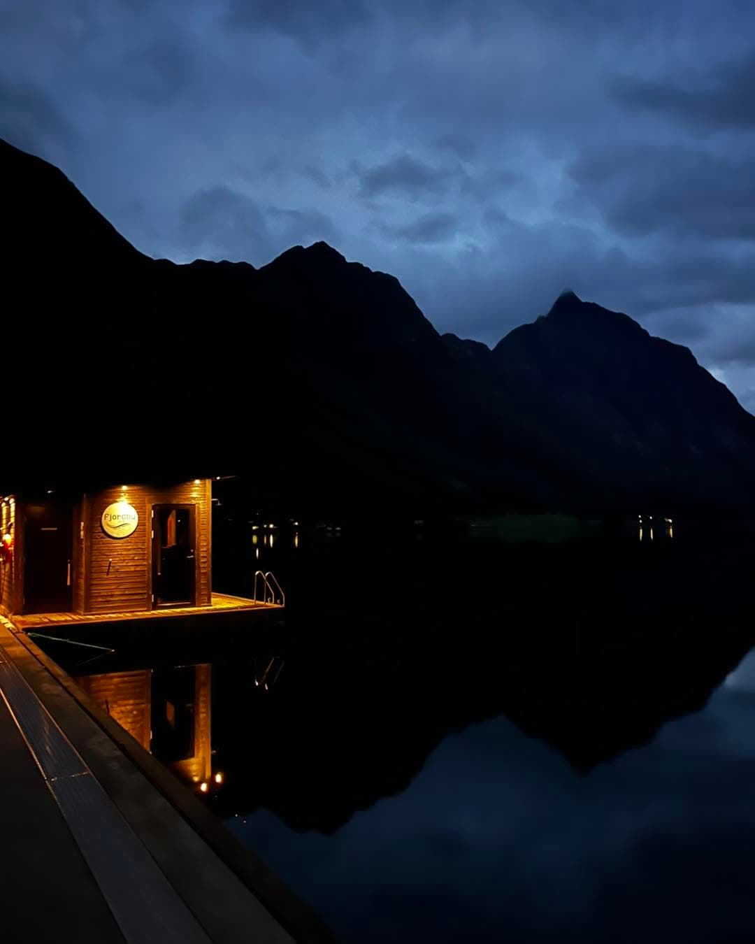 Badstua opplyst i blåtimen, med varmt lys frå vindauga og fjella som mørke silhuettar i spegelblank fjord.