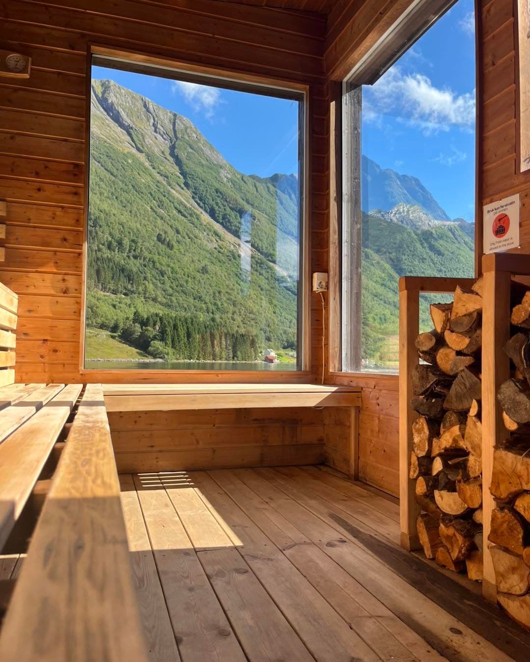 Inne i badstua: benkar i lyst tre, to store vindauge ut mot fjell og fjord, og opplagd ved ved veggen.